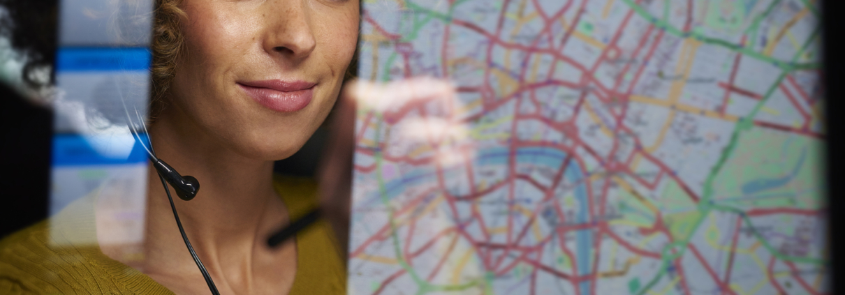 A young woman with a curious expression stands in front of a map, attentively examining it with her finger tracing a route. She appears engaged and focused, ready to explore and navigate the world