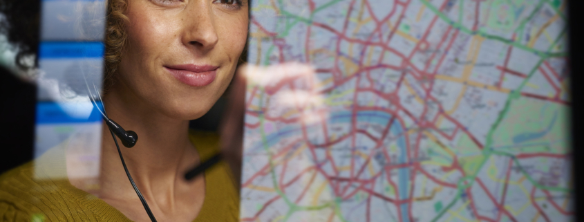 A young woman with a curious expression stands in front of a map, attentively examining it with her finger tracing a route. She appears engaged and focused, ready to explore and navigate the world